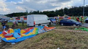 na zdjęciu miejsce zdarzenia, gdzie na trawie leżąca na ziemi dmuchana atrakcja, obok biały dostawczy samochód, obok niego inne samochody, osoby, strażacy, ratownicy, policjanci, w oddali widoczny helikopter na łące