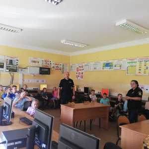 na zdjęciu dzielnicowy stoi na środku sali lekcyjnej, obok strażniczka miejska, wokół nich dzieci przy biurkach szkolnych