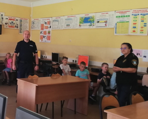 na zdjęciu dzielnicowy stoi na środku sali lekcyjnej, obok strażniczka miejska, wokół nich dzieci przy biurkach szkolnych