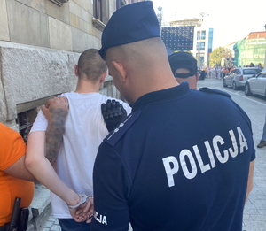 na zdjęciu policjant kuje w kajdanki zatrzymanego, którego trzyma ochrona