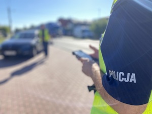 na zdjęciu fragment policyjnego munduru, w oddali inny policjant przy kontrolowanym czarnych samochodzie