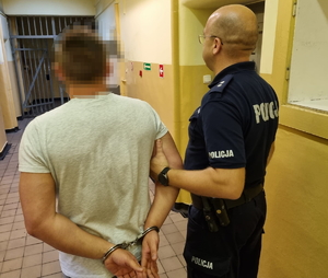 na zdjęciu policjant trzyma za rękę zatrzymanego w korytarzu komendy