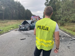 na zdjęciu policjant w odblaskowej kamizelce stoi na jezdni, a przed nim rozbity pojazd