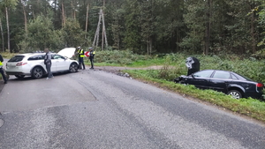 na zdjęciu miejsce zdarzenia gdzie stoi pojazd, obok niego policjanci i inne osoby, drugi samochód w rowie po prawej stronie zdjęcia