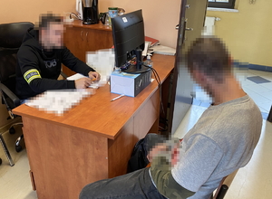 na zdjęciu policjant przy biurku z opaską z napisem policja na ramieniu, przed nim zatrzymany siedzi na krześle
