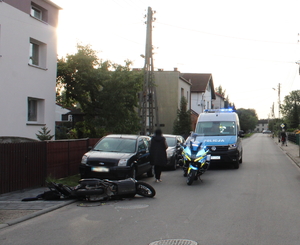 na zdjęciu przewrócony motocykl na jezdni, za którym stoją dwa samochody osobowe, przy których stoi osoba i policyjny radiowóz i motocykle