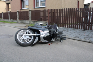 na zdjęciu uszkodzony motocykl leżący na asfalcie, za nim płot i budynek