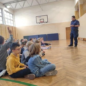 na zdjęciu policjant przed dziećmi siedzącymi na parkiecie w sali gimnastycznej