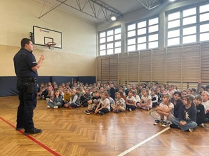 na zdjęciu policjant przed dziećmi siedzącymi na parkiecie w sali gimnastycznej