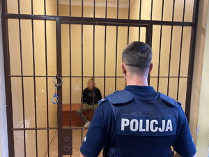 na zdjęciu zatrzymany w policyjnej celi, przed kratą tyłem stoi policjant w mundurze