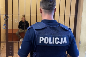 na zdjęciu zatrzymany w policyjnej celi, przed kratą tyłem stoi policjant w mundurze