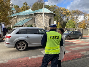na zdjęciu policjant rozmawiający z osoby przy przejściu dla pieszych, na jezdni widoczne inne pojazdy i osoby, w tle płot i cmentarz
