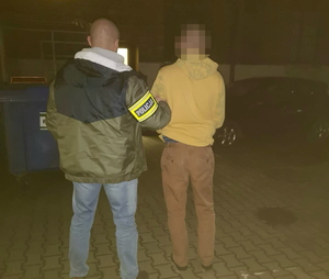 na zdjęciu policjant w ubraniu cywilnym z opaską na ramieniu z napisem policja, trzyma za rękę zatrzymanego