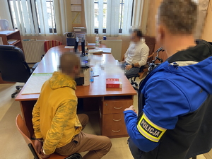 na zdjęciu policjant z opaską na ramieniu, przed nim na krześle przy biurka zatrzymany w kajdankach i policjant