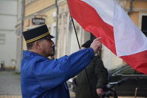 na zdjęciu dyrygent orkiestry, obok polska flaga