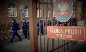 na zdjęciu fragment ogrodzenia na którym widoczne godło i napis szkoła policji w słupsku, za ogrodzeniem maszerujący policjanci