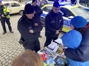 na zdjęciu policjanci przy stoliku z innymi osobami, za nimi radiowóz z włączonymi niebieskimi sygnałami