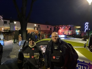 na zdjęciu policjant ze strażakiem przy radiowozie, za nimi scena na małym rynku w Kaletach i inne osoby