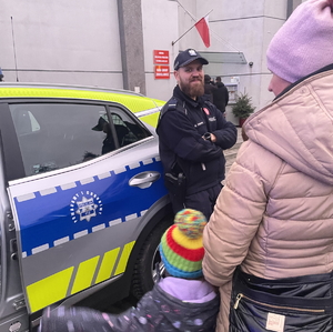 na zdjęciu policjant oparty o radiowóz z uśmiechem na twarzy, przed nim kilka osób