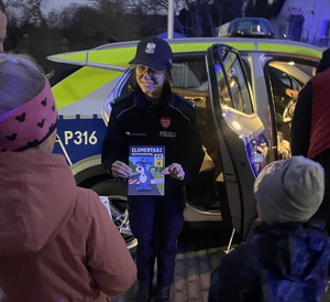 na zdjęciu policjantka z elementarzem w ręku, za nią radiowóz, a przed nią osoby