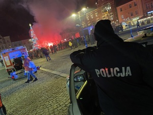 na zdjęciu policyjny radiowóz, a przy nim policjant, przed którym na płycie rynku odpalane są środki pirotechniczne koloru czerwonego, wokół stoją ludzie