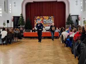 na zdjęciu policjant w sali widowiskowej w trakcie rozmów z wolontariuszami na temat bezpieczeństwa, którzy siedzą po bokach sali na krzesłach