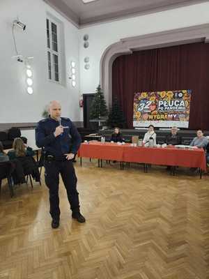 na zdjęciu policjant w sali widowiskowej w trakcie rozmów z wolontariuszami na temat bezpieczeństwa, którzy siedzą po bokach sali na krzesłach