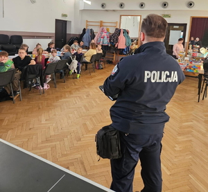 na zdjęciu dzielnicowy przed dziećmi, które siedzą na krzesłach przy stołach w sali