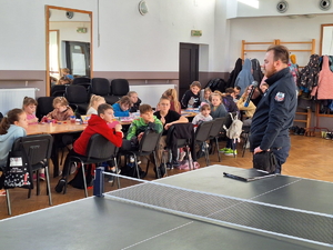 na zdjęciu dzielnicowy przed dziećmi, które siedzą na krzesłach przy stołach w sali