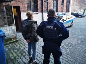 na zdjęciu policjant, obok niego zatrzymana, przed nimi radiowozy policyjne