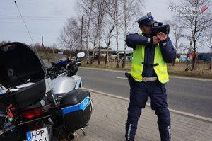 na zdjęciu policjant przy motocyklu policyjnym, trzymający w rękach urządzenie do pomiaru prędkości
