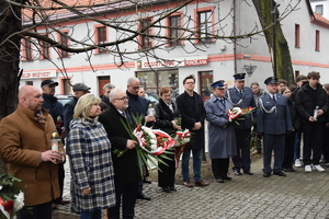 na zdjęciu osoby zgromadzone przed tablicą, z kwiatami w rękach oraz zniczami stoi komendant policji dyrektor aresztu, przedstawiciele władz miasta i powiatu oraz inne osoby