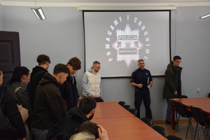 na zdjęciu policjant w sali, a obok niego wchodzący do sali i siadający na krzesłach za stołami, za policjantem logo policji z napisem pomagamy i chronimy policja tarnowskie góry