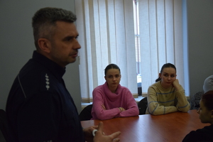 na zdjęciu policjant profilem do aparatu, za nim przy stole uczennice