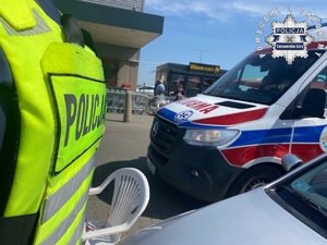 na zdjęciu tylna część kamizelki odblaskowej policjanta, obok karetka pogotowia, ludzie i budynek sklepu