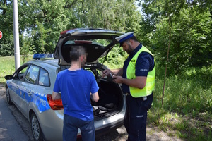 na zdjęciu policjant z mężczyzną przy radiowozie, w którym otwarta jest klapa bagażnika