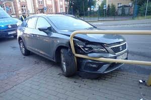 na zdjęciu samochód osobowy wbity a barierkę chodnikową, za nim idący policjant oraz fragment radiowozu
