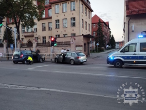 na zdjęciu miejsce zdarzenia, od prawej radiowóz na jezdni, następnie jasny samochód osobowy wbity w barierkę chodnikową, obok kolejny w poprzek drogi, przy którym policjant, za pojazdami inne osoby oraz budynki