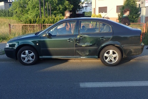 na zdjęciu ciemny samochód osobowy z uszkodzonym bokiem zaparkowany na chodniku, za nim osoby