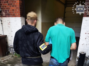 na zdjęciu policjant w ubiorze cywilnym z opaską na ręku z napisem policja prowadzi zatrzymanego w kajdankach