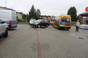 na zdjęciu miejsce wypadku, gdzie widać policyjny radiowóz, karetkę pogotowia, a pomiędzy nimi czarny samochód osobowy, po prawej stronie budynek sklepu, za pojazdami inne zaparkowane pojazdy