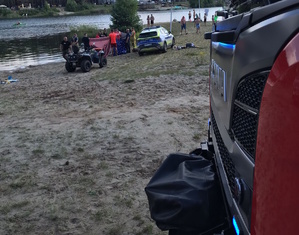 na zdjęciu fragment wozu strażackiego, w oddali na plaży radiowóz, parawan i ratownicy