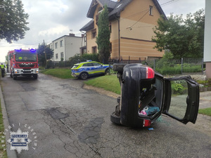 na zdjęciu czarny samochód osobowy na jezdni na boku, obok wóz strażacki, strażacy i policjanci