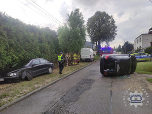 na zdjęciu czarny samochód osobowy na jezdni przewrócony na bok, obok policjant, a po lewej stronie policjanta zaparkowany czarny samochód osobowy z uszkodzonym przodem. przed policjantem strażacy