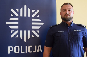 Na zdjęciu dzielnicowy Moj przy banerze z napisem policja