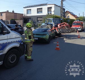 na zdjęciu fragment radiowozu, przy którym stoi strażak, przed nim samochód, który uderzył w traktor, za tymi pojazdami wóz strażacki