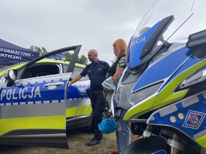 na zdjęciu policyjny motocykl i radiowóz a pomiędzy nimi policjant z osobami