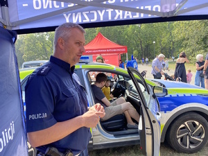 na zdjęciu policjant pod namiotem informacyjnym, a za nim uczestnicy pikniku w radiowozie