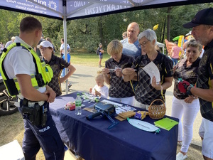 na zdjęciu policjant w namiocie informacyjnym przy stole z upominkami, przy którym stoją inne osoby