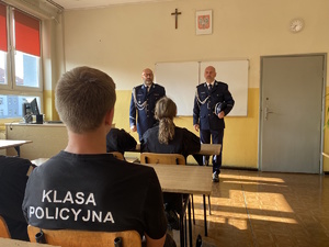 na zdjęciu w pierwszym planie uczeń w ławce z koszulką z napisem klasa policyjna, a przed nim stojący policjanci